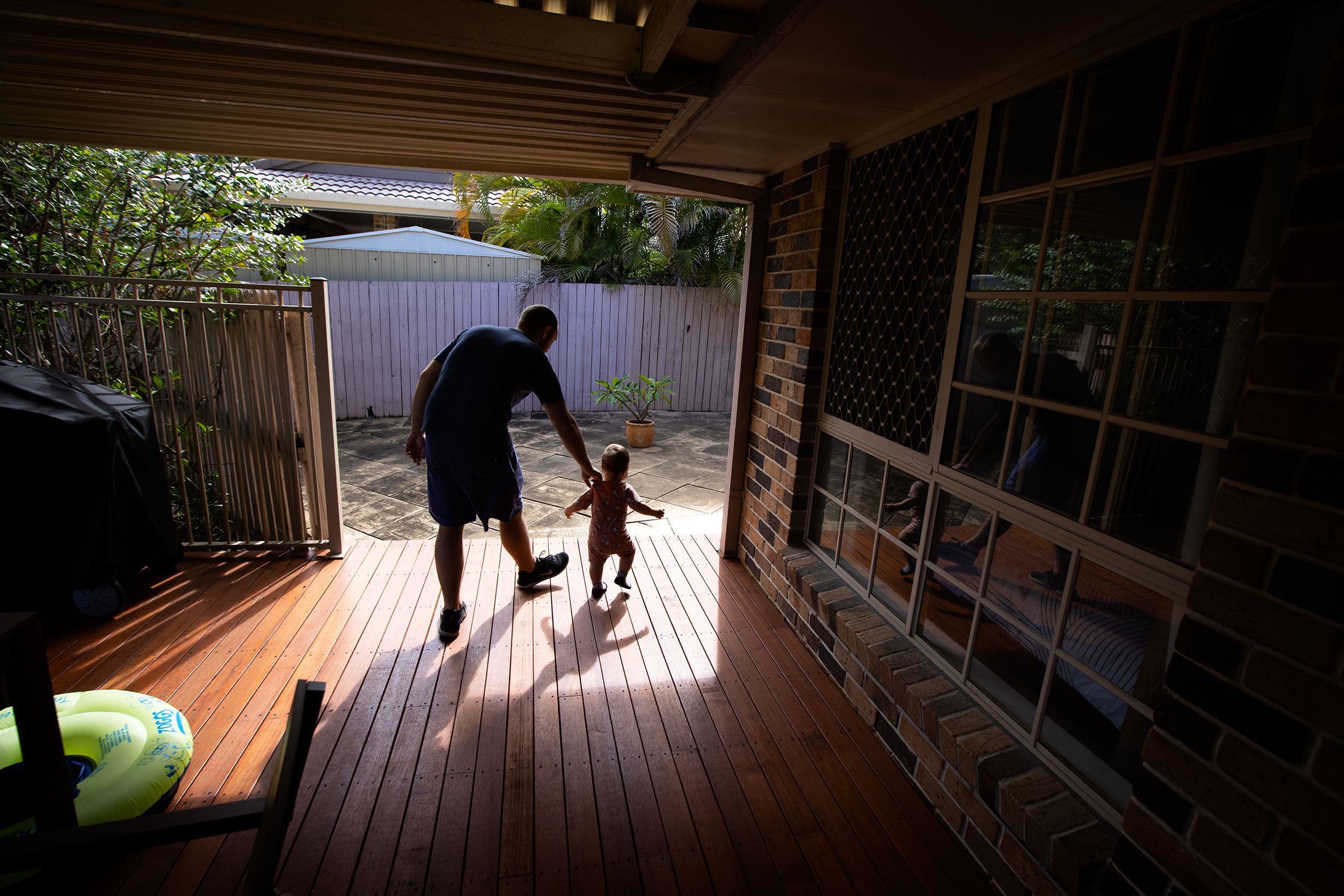 A man holds a toddler as they walk from a covered deck onto a sunlit paved square