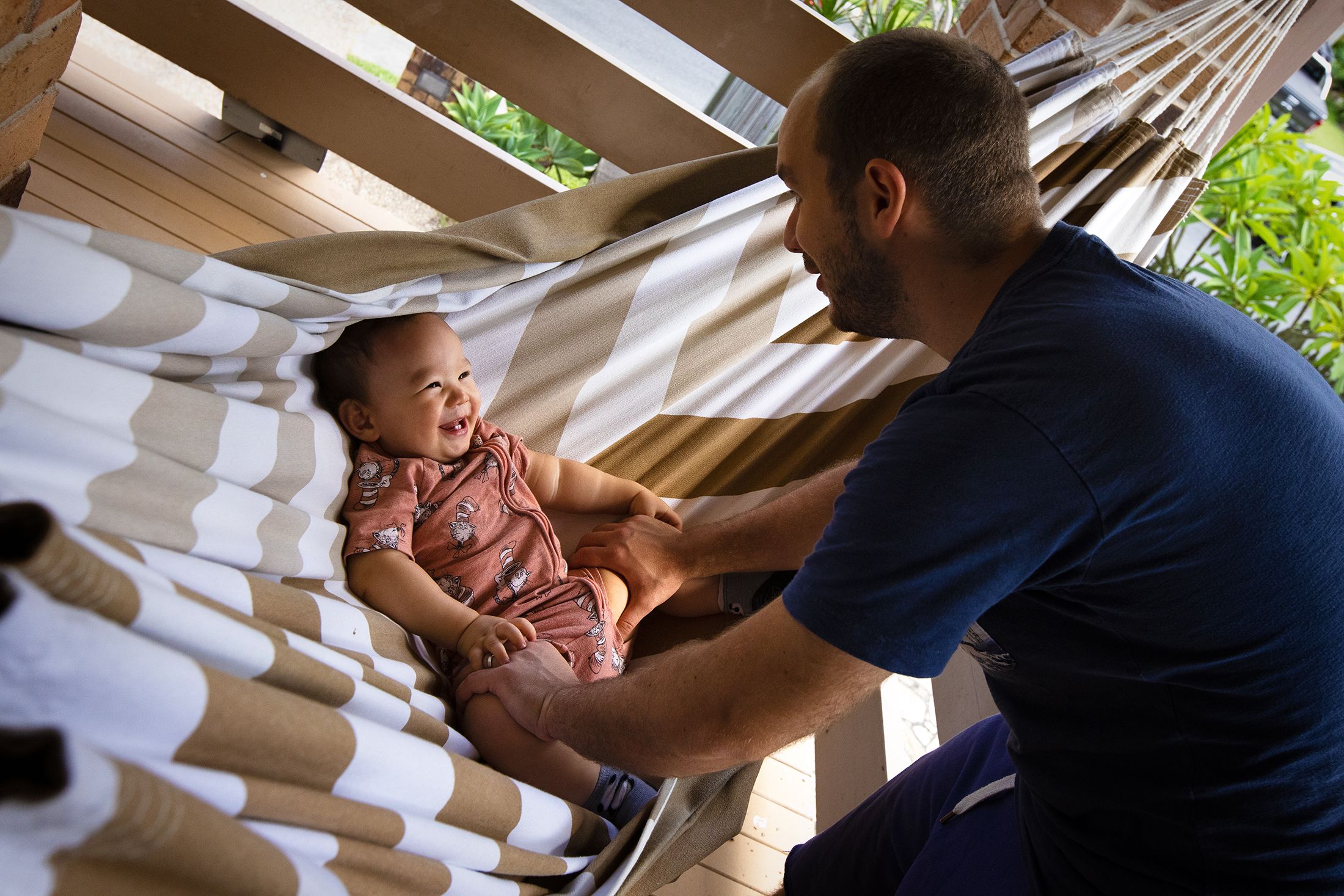 A man rocks his toddler in a hammock as the child laughs. Both are looking at each other happily.
