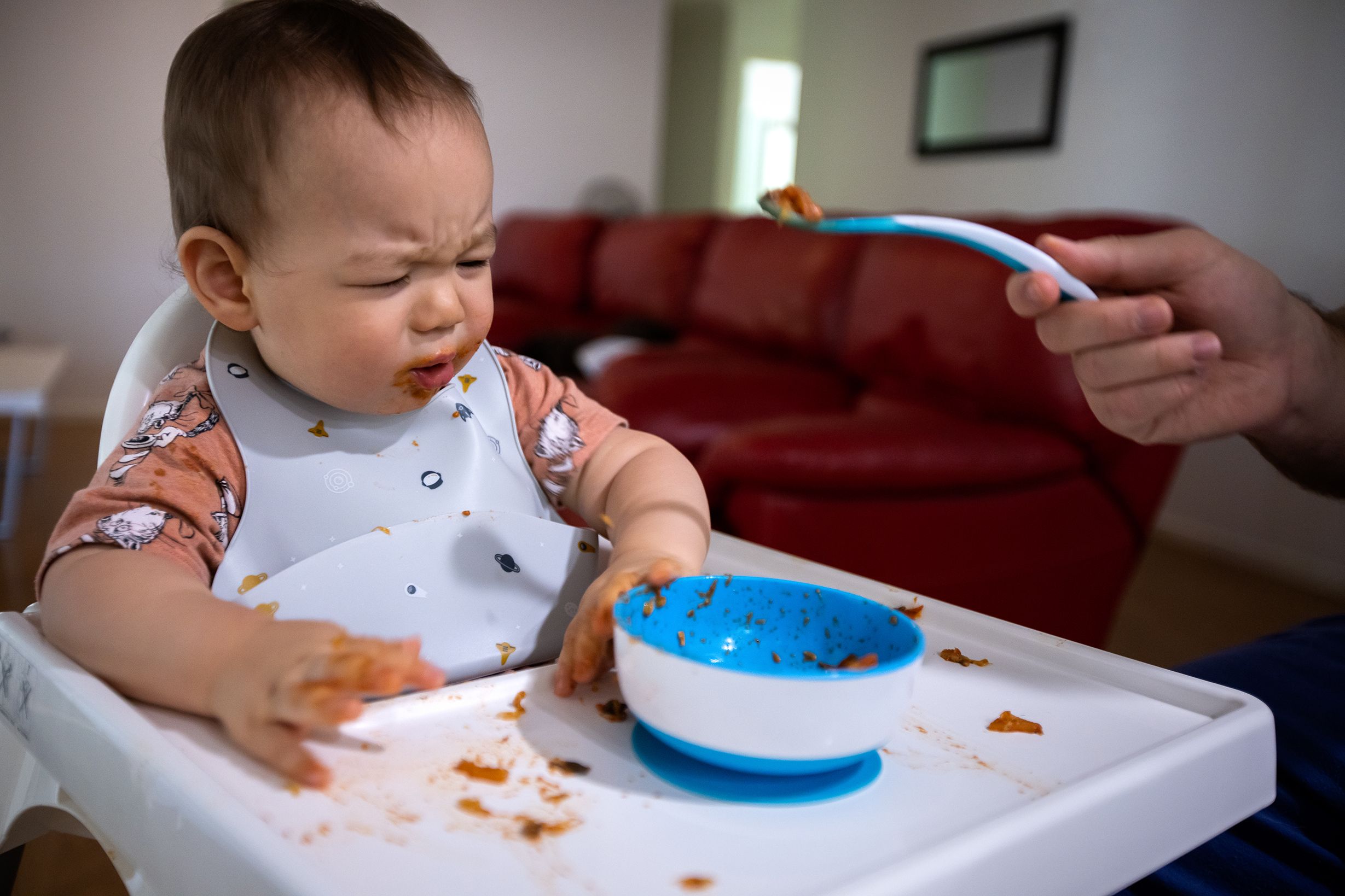 A male toddler sits in a high chair. A hand feeds him food on a spoon. There is food debris on the chair.