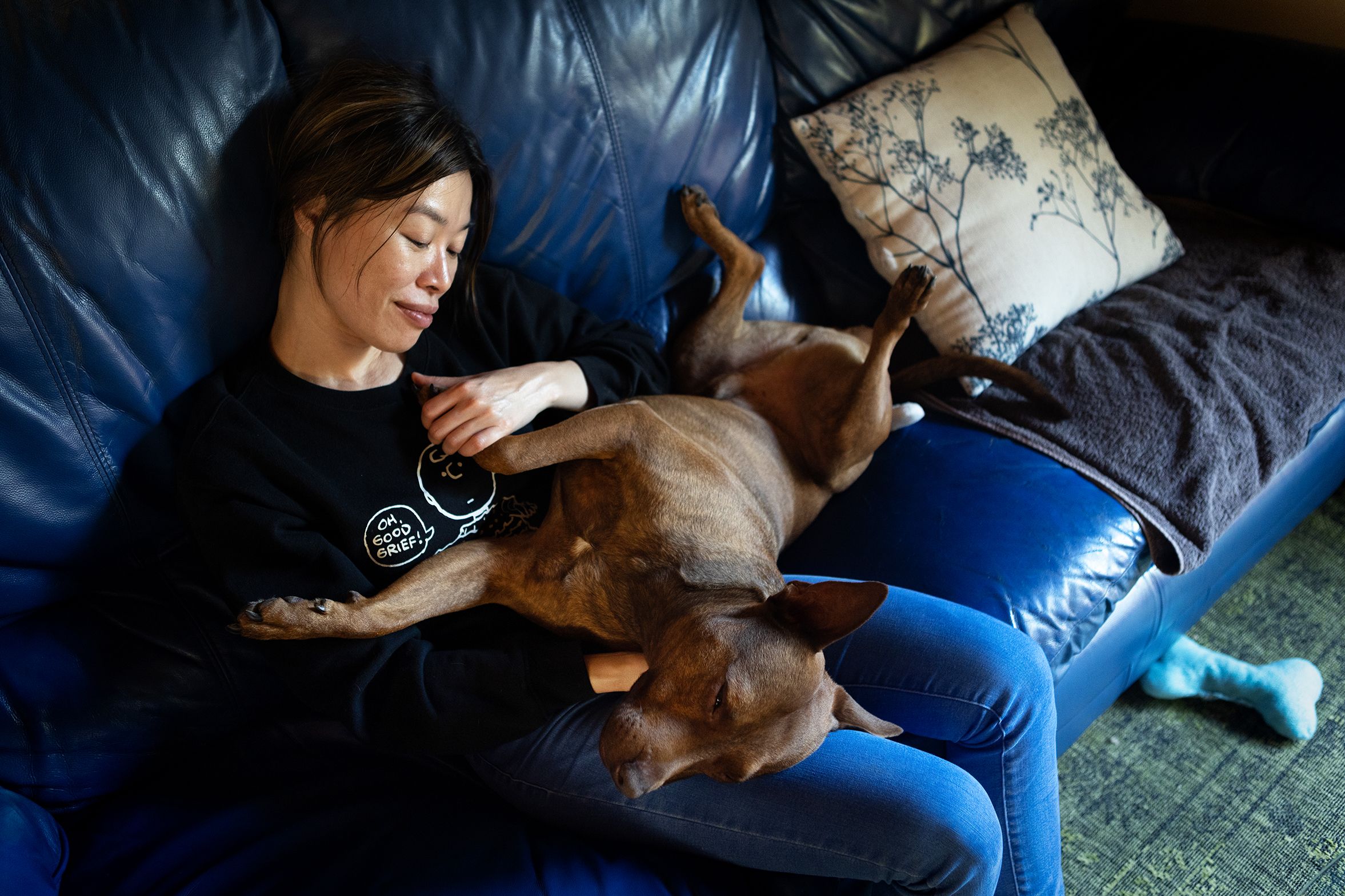 A Vietnamese woman with long black hair holds her dog as they sit on a blue leather sofa in a living room.