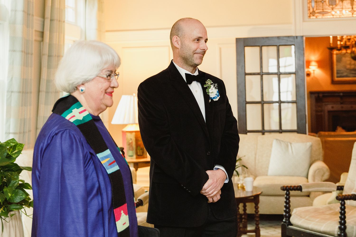 Wedding ceremony attendees stand in formal attire against a cream-colored wall with elegant lighting fixtures.