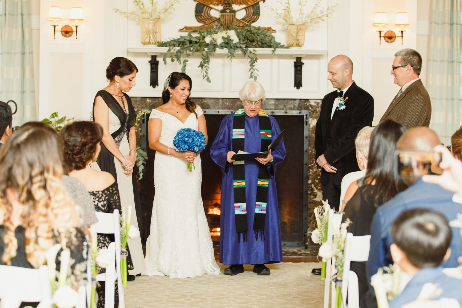 Wedding ceremony guests and wedding party members gather in an elegant indoor venue with decorative greenery.