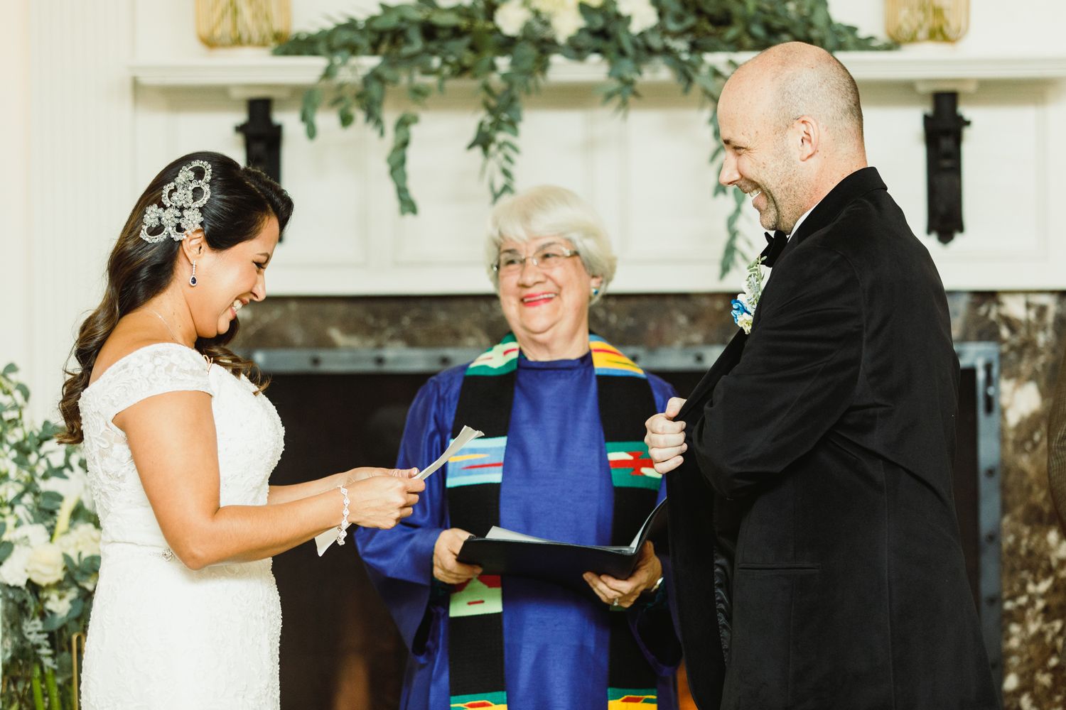 Wedding ceremony participants exchange vows in front of decorative greenery and floral arrangements.