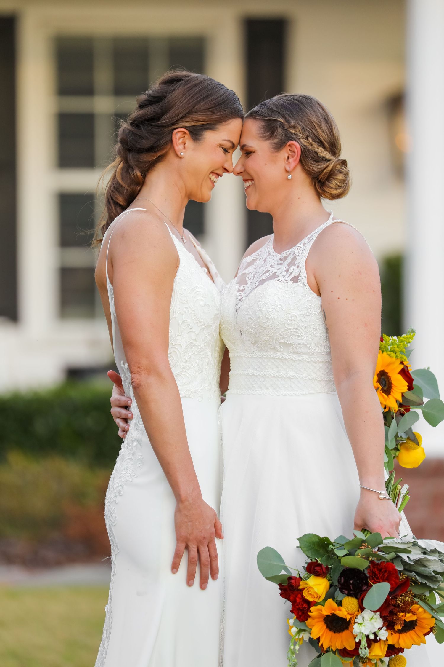 Two brides in white lace dresses share an intimate moment while holding sunflower bouquets.
