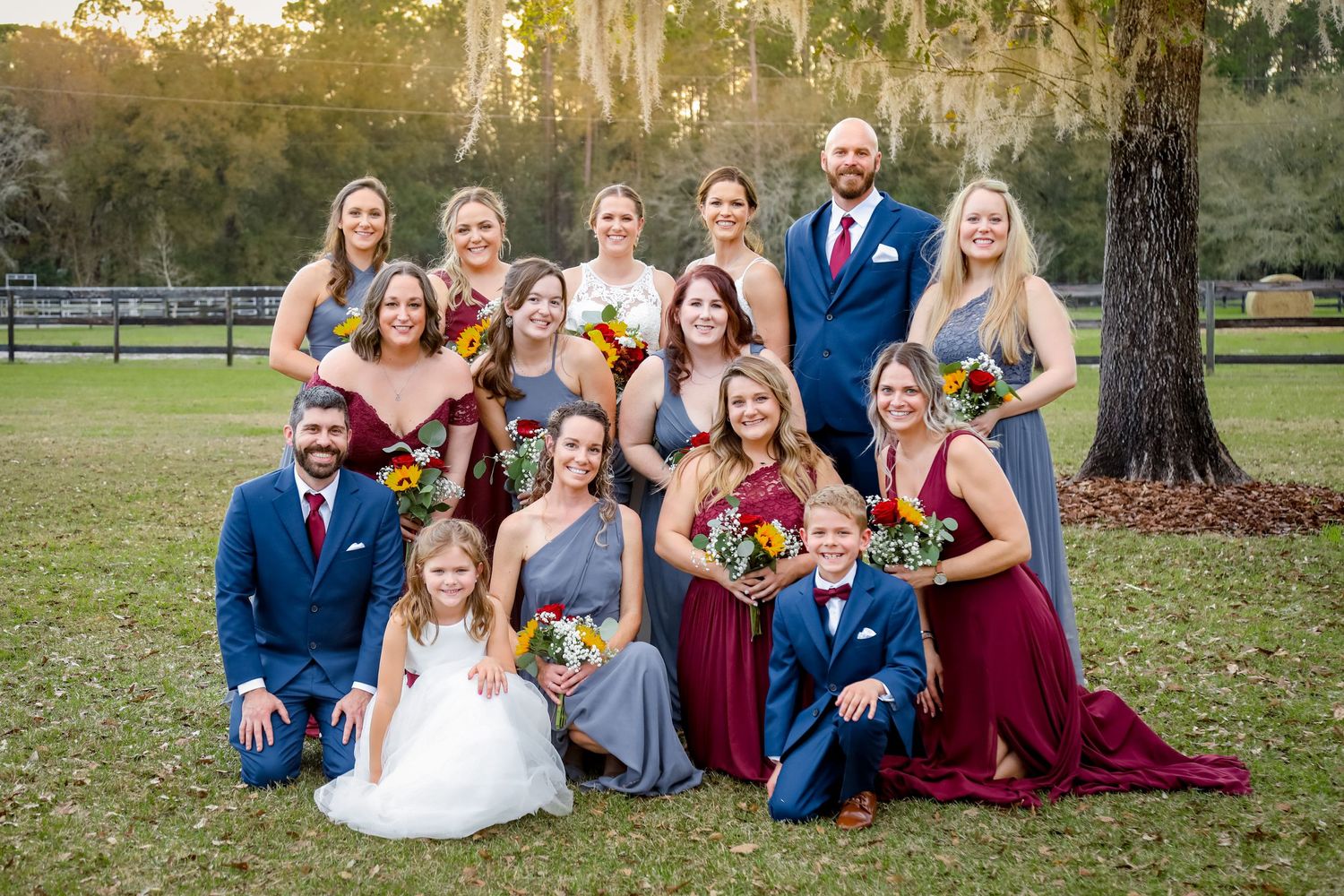 Wedding party wearing burgundy and blue attire poses together outdoors on grass under trees at sunset.