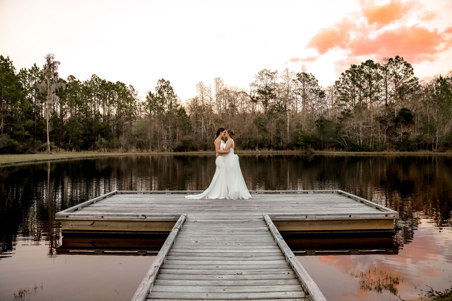 Wedding photo of person in white dress standing on wooden dock over lake at sunset.