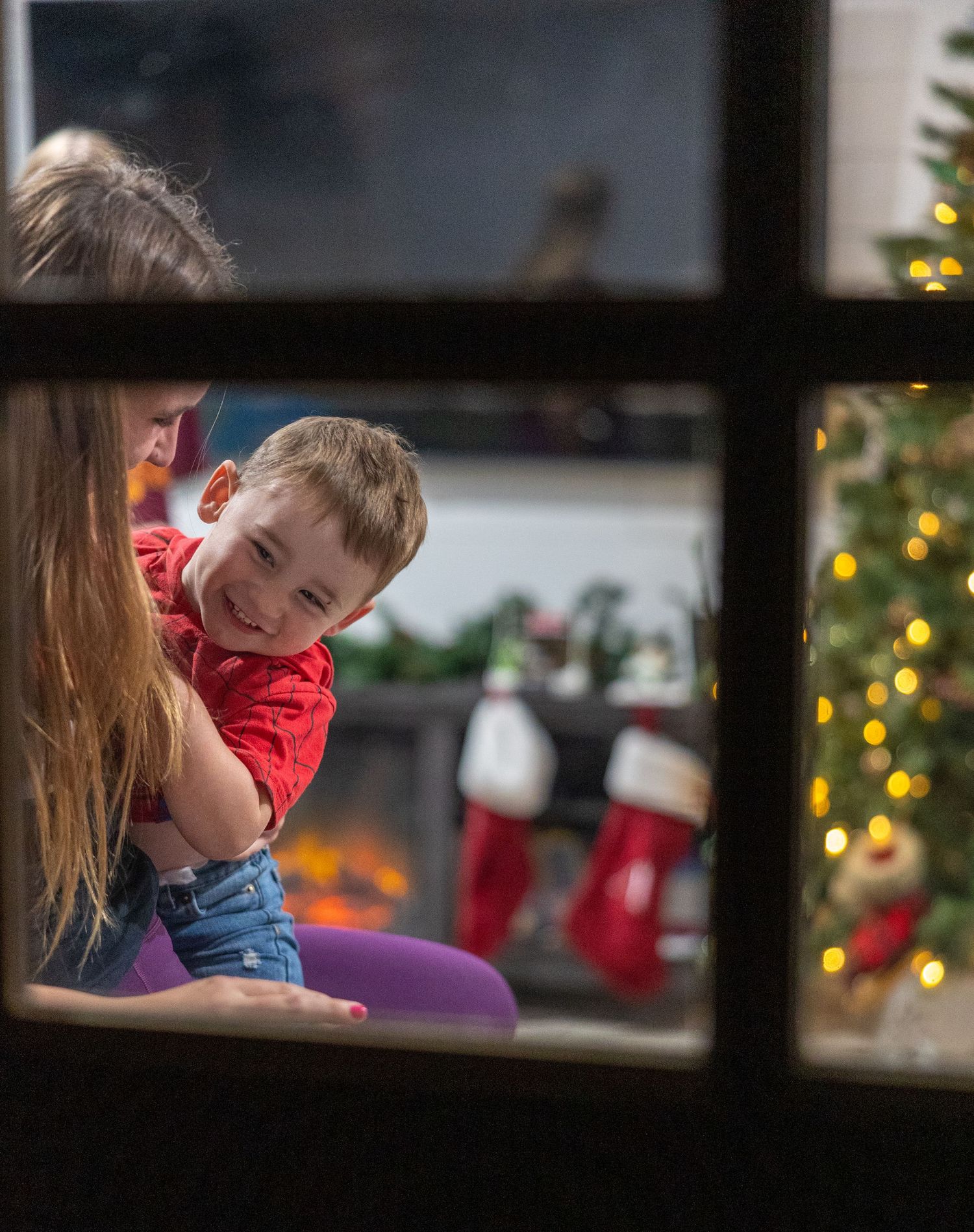 A decorated Christmas tree and stockings visible through a window frame in warm evening light.