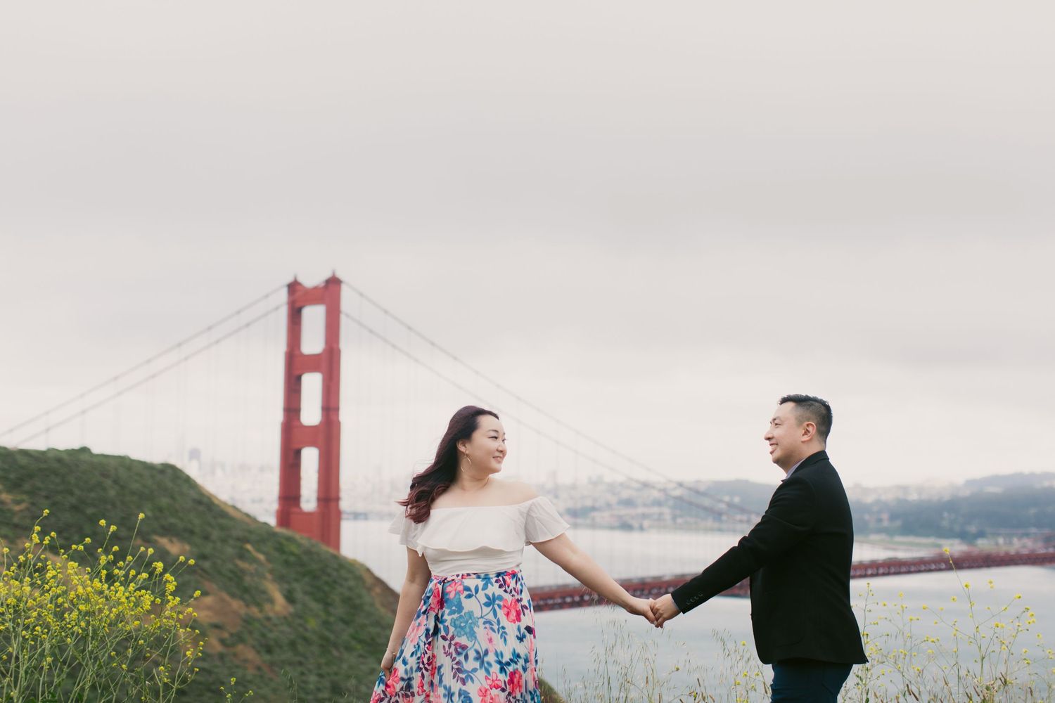 Couple holding hands near Golden Gate Bridge with yellow wildflowers in foreground.