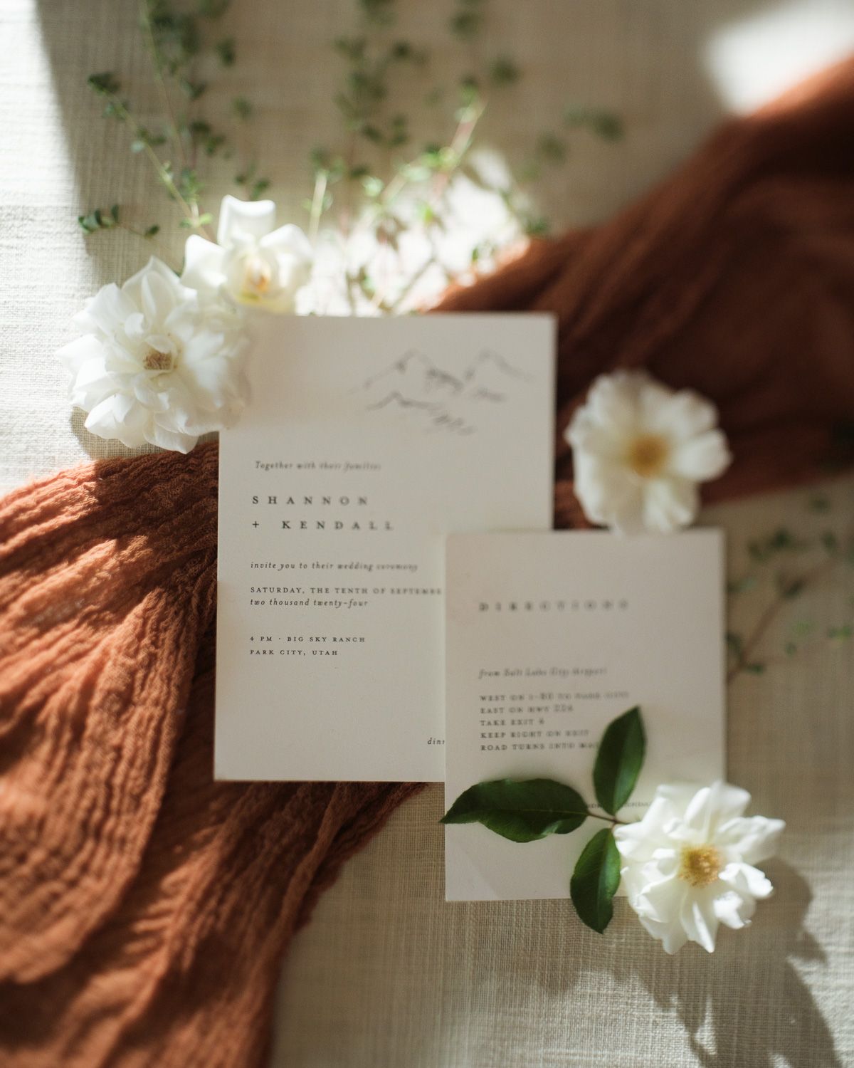 White minimalist wedding invitation cards with delicate white flowers and green leaves against a textured rust-colored fabric backdrop.