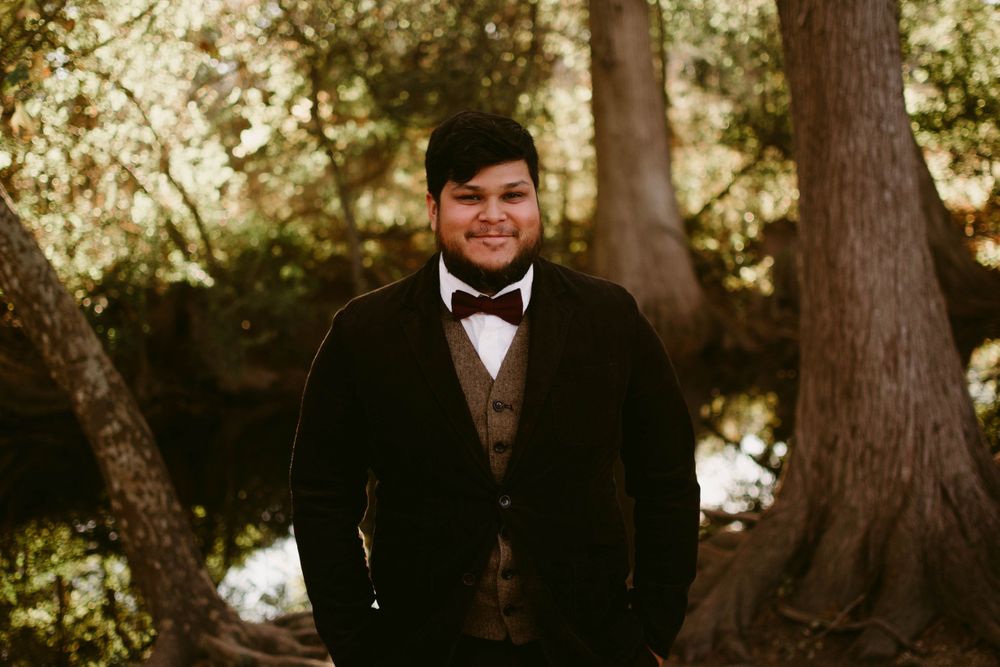 A formal portrait in a wooded setting featuring someone in a black tuxedo and bowtie.