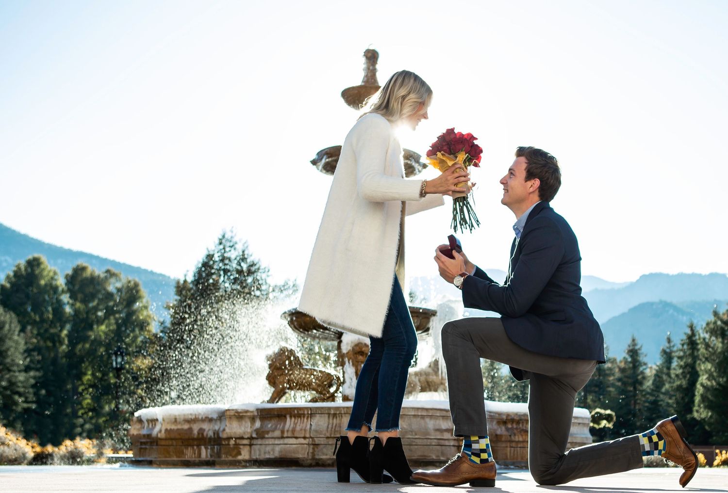A surprise proposal moment captured near a fountain with mountain views in the background.