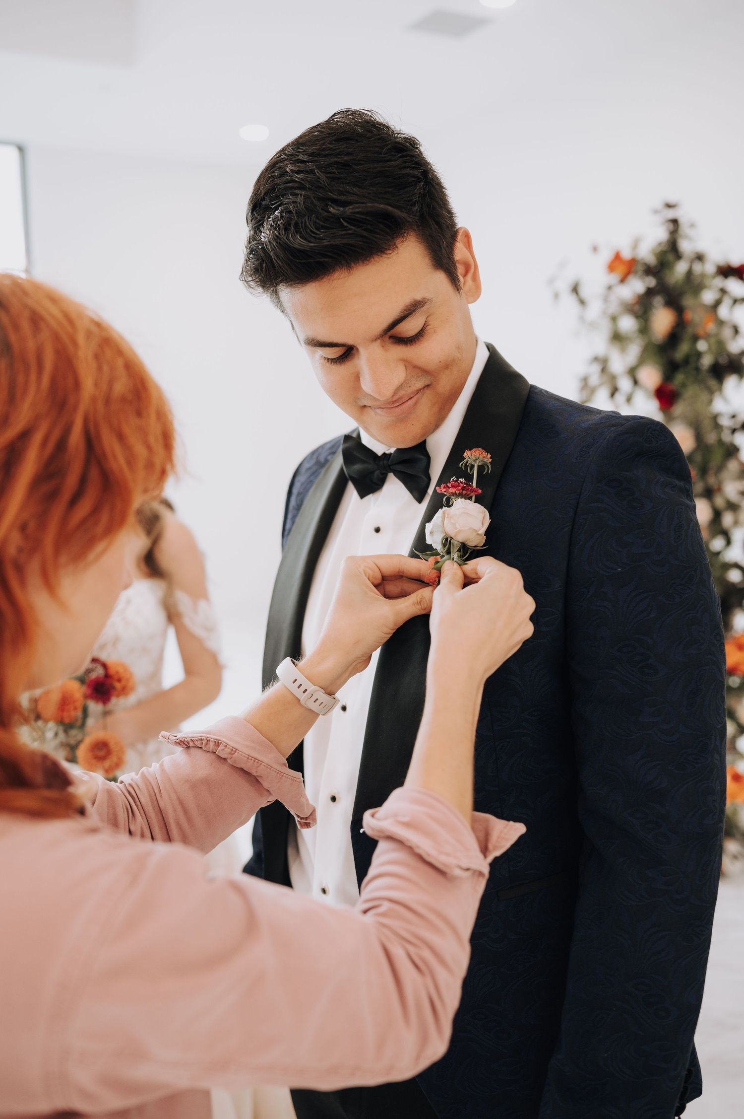 A boutonniere being pinned onto a formal black tuxedo with floral decor in background.