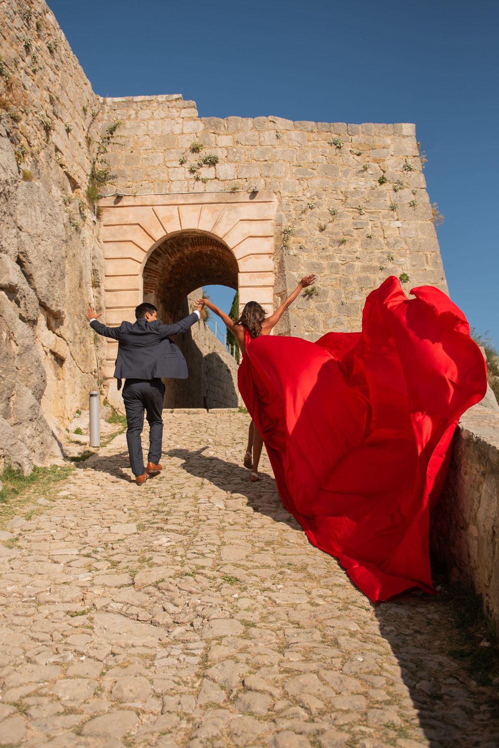 Two figures stand near an ancient stone archway with a flowing red dress billowing in the wind.