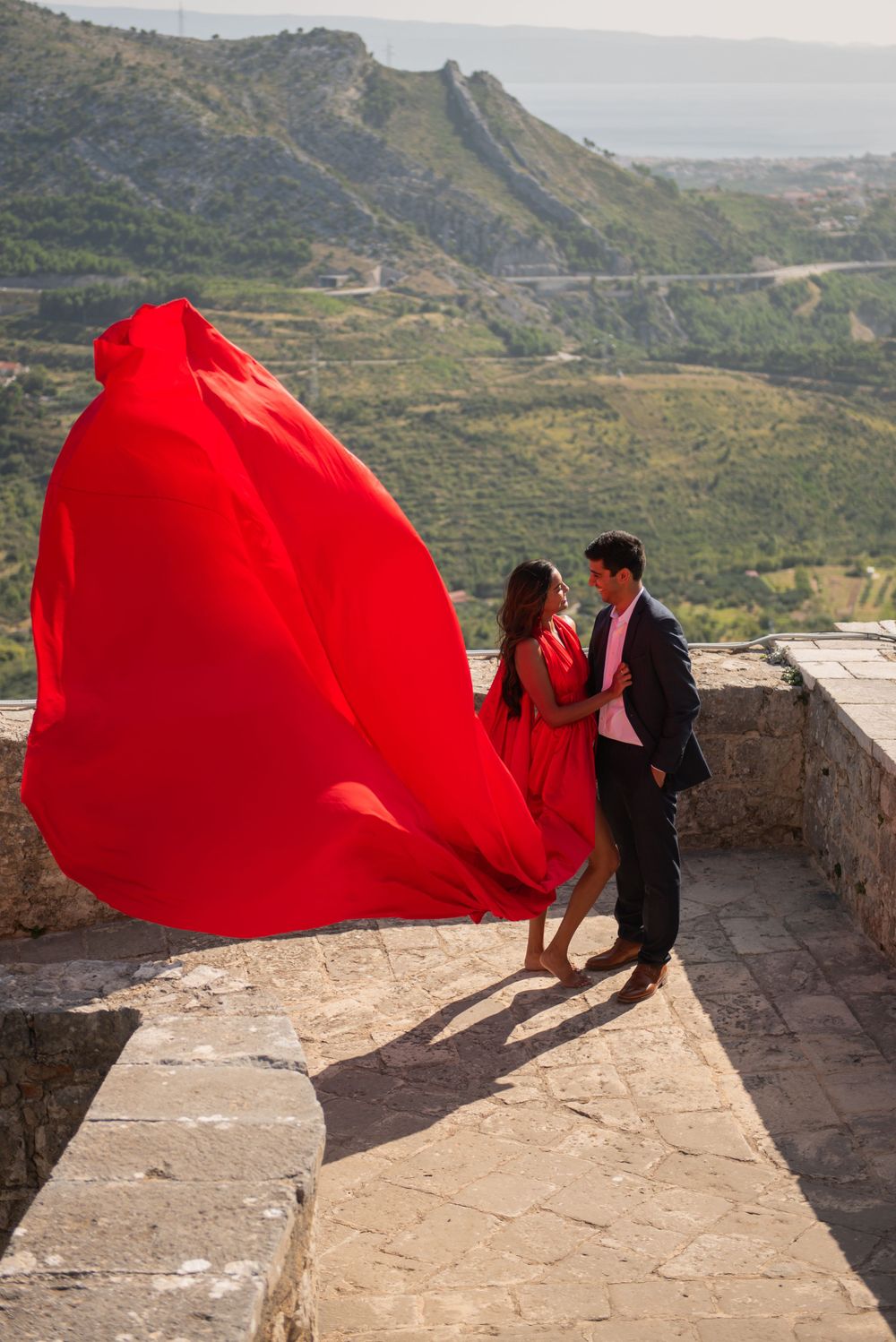 A romantic couple stands on a scenic overlook with a flowing red fabric dramatically billowing in the wind.