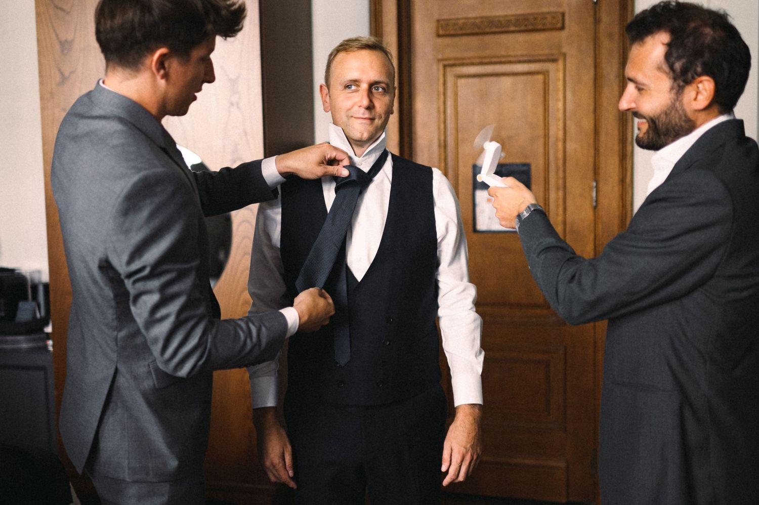 Three men in formal attire interacting in front of a wooden door during a celebratory event.