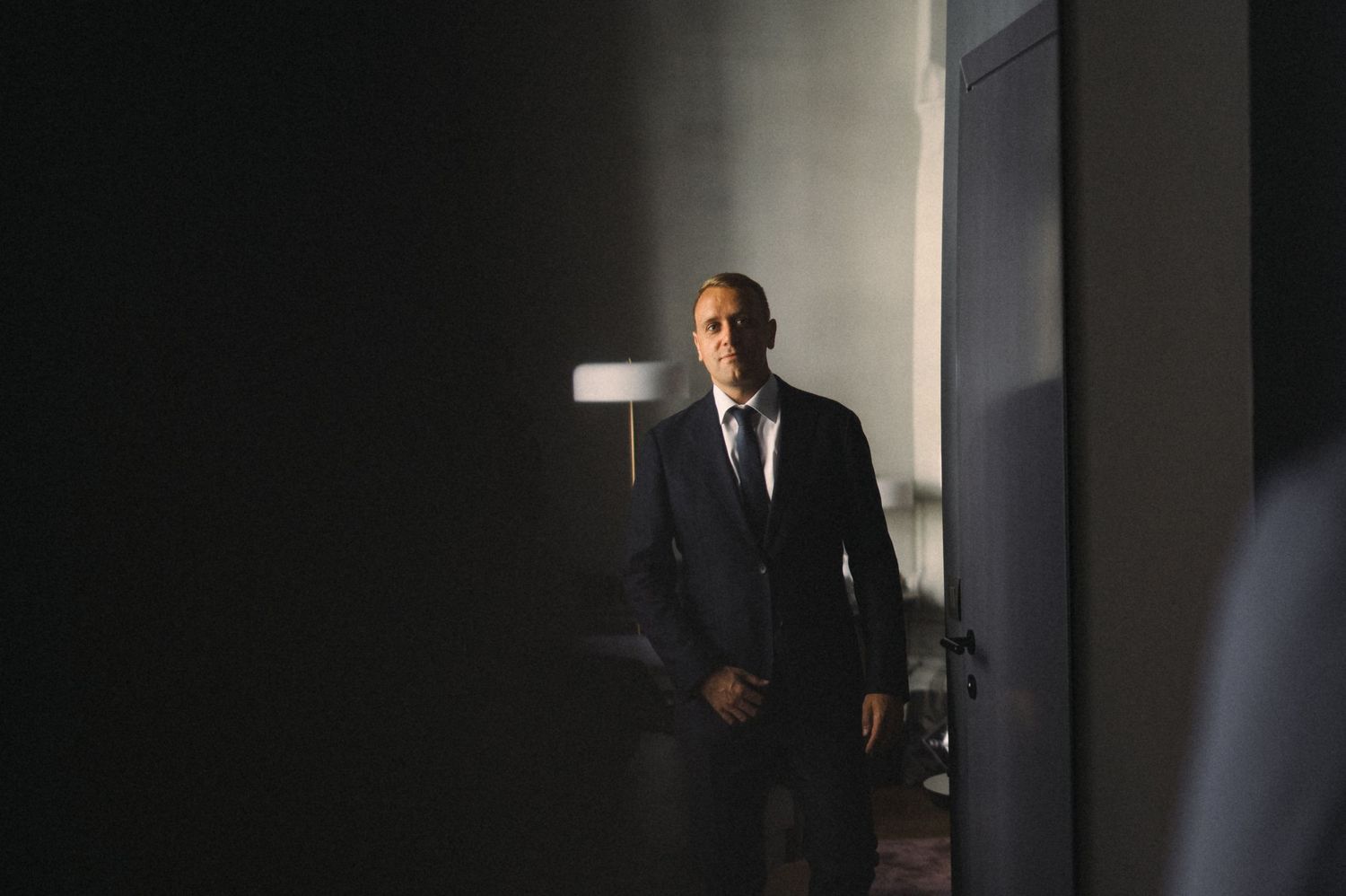 A dramatic low-light portrait of a person in a dark suit standing in a shadowy doorway.