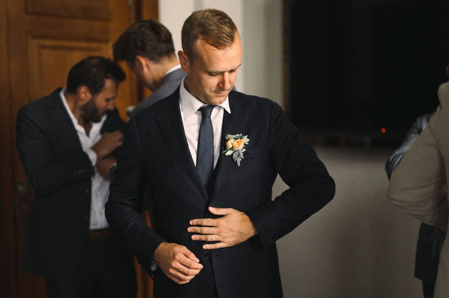 Groom in classic black tuxedo preparing for wedding ceremony with groomsmen in background.