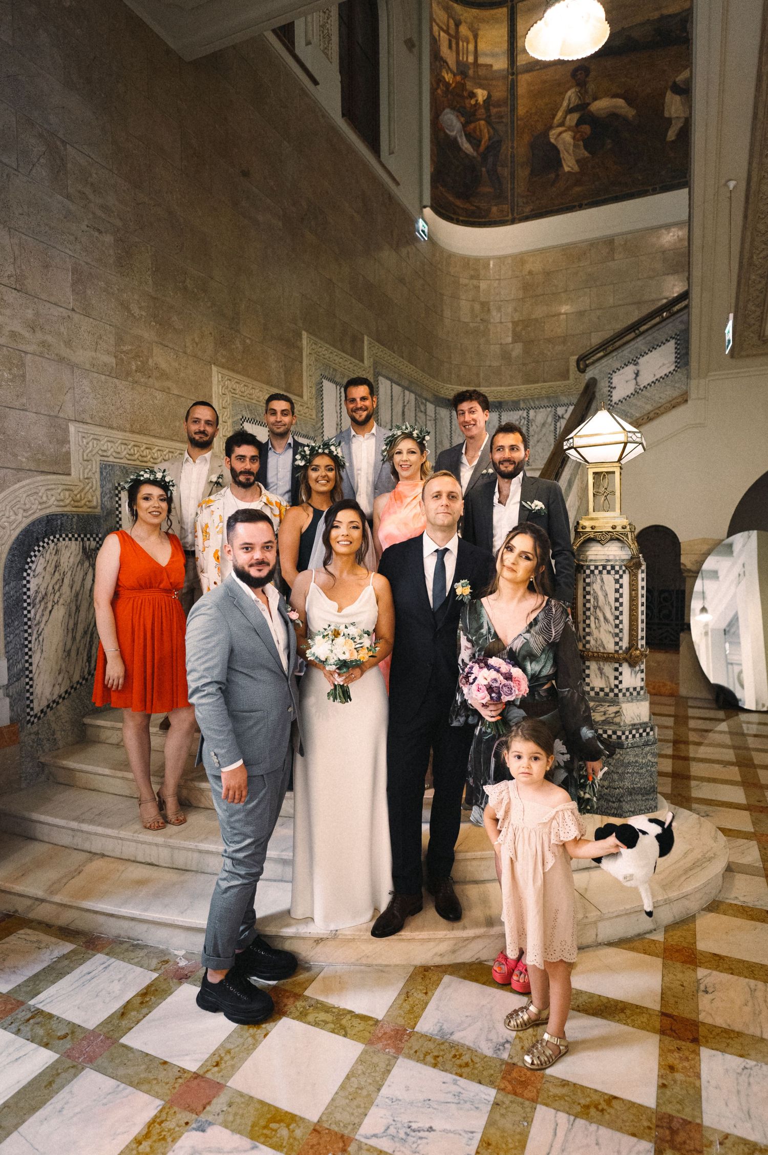 A wedding party poses on an elegant marble staircase with ornate murals decorating the walls and ceiling.