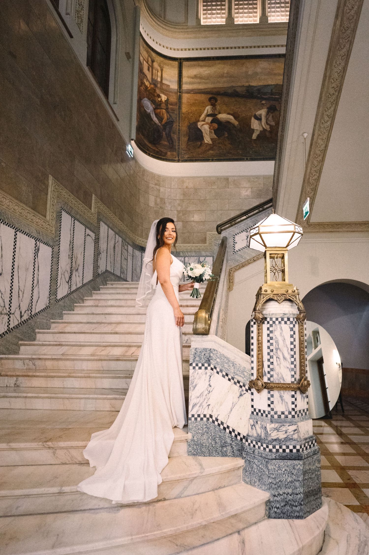 A bride in a flowing white dress poses on a grand marble staircase with decorative lanterns and wall murals.