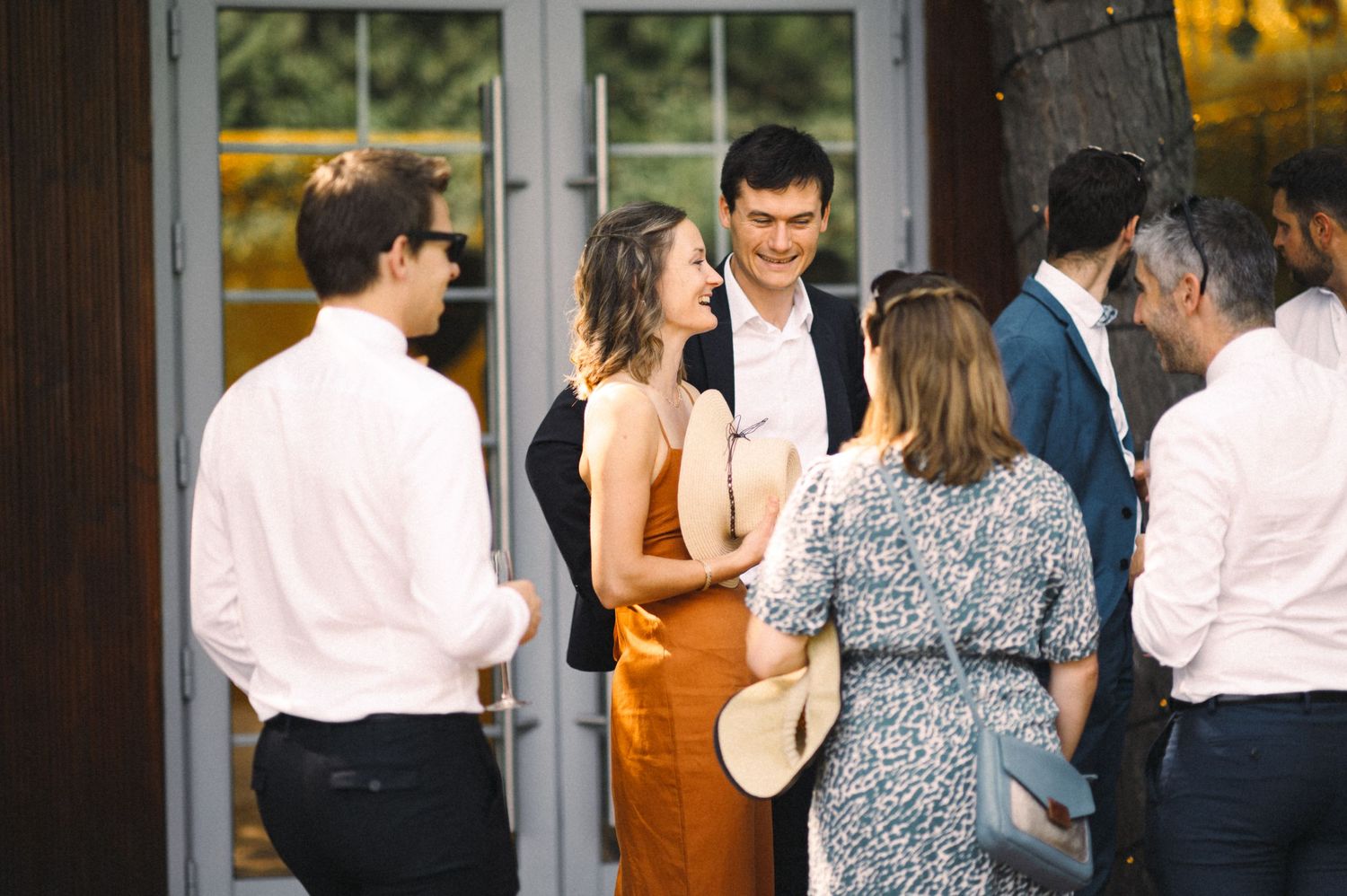 Wedding guests mingle at an outdoor reception gathering near glass doors.