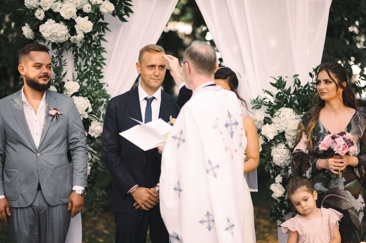 Wedding ceremony in progress under decorated arch with officiant in white robes.