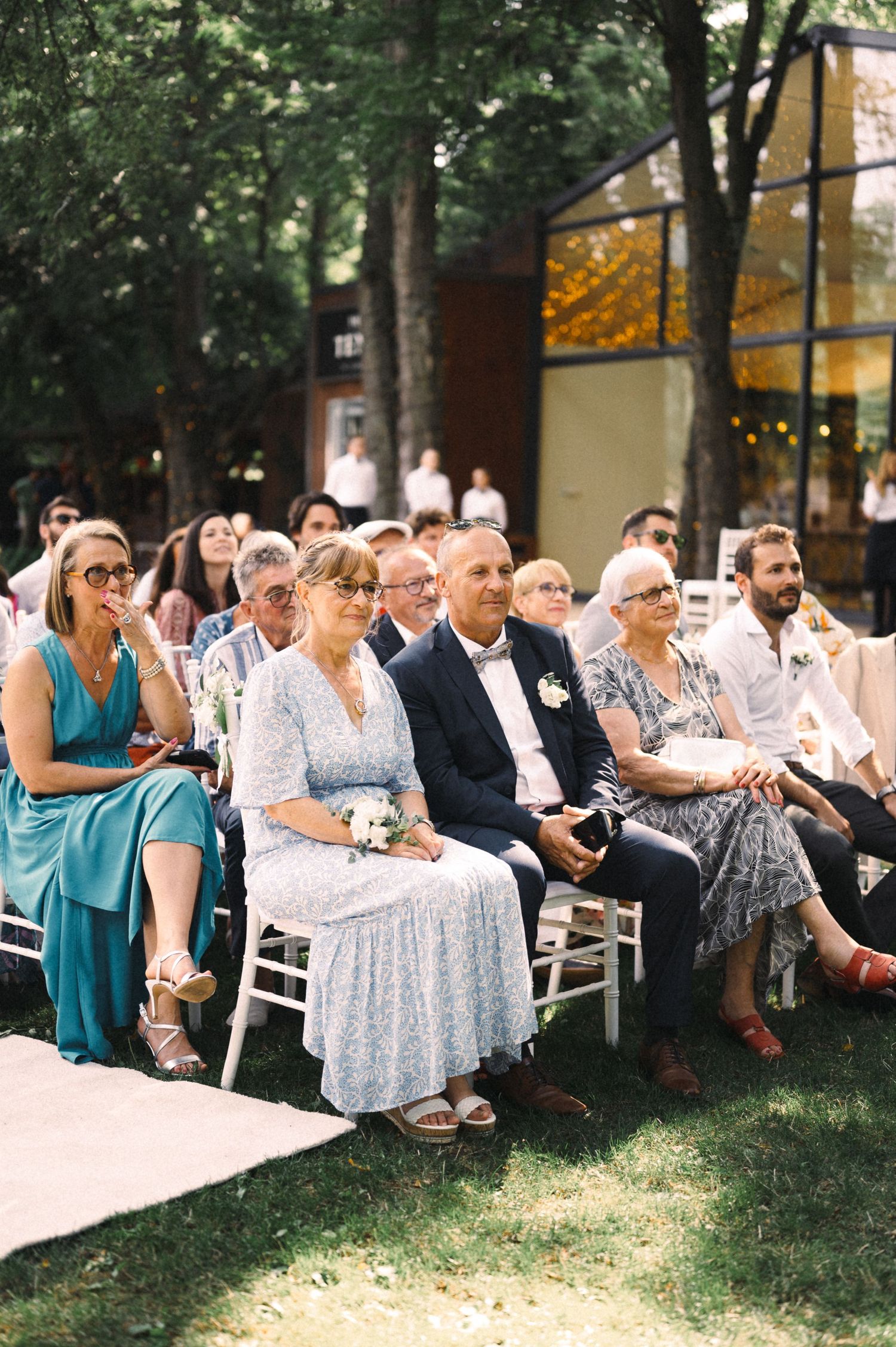 Wedding guests seated on white chairs during outdoor garden ceremony.