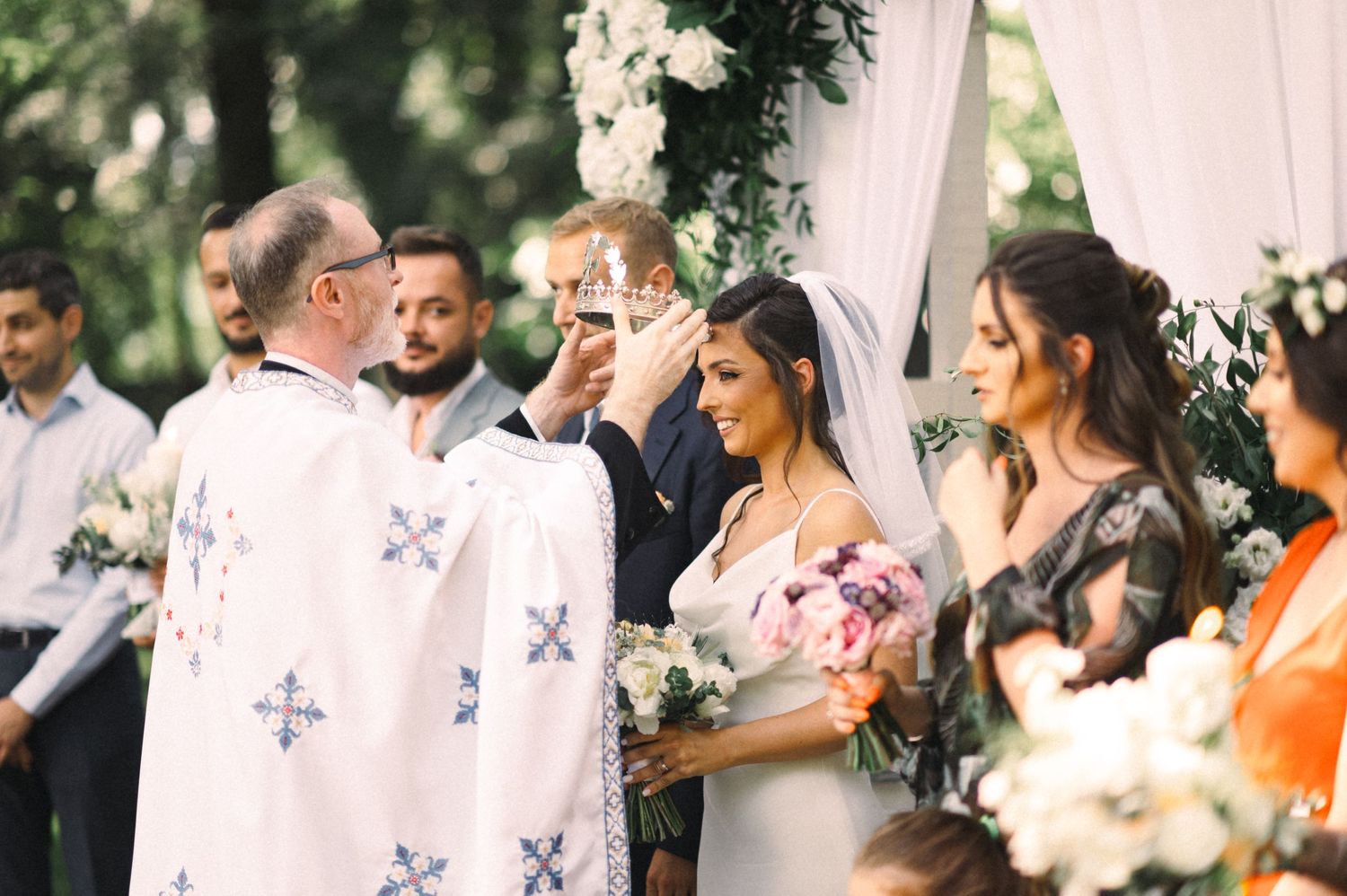 Intimate wedding ceremony under pink arch with clergy in white robes adorned with crosses.