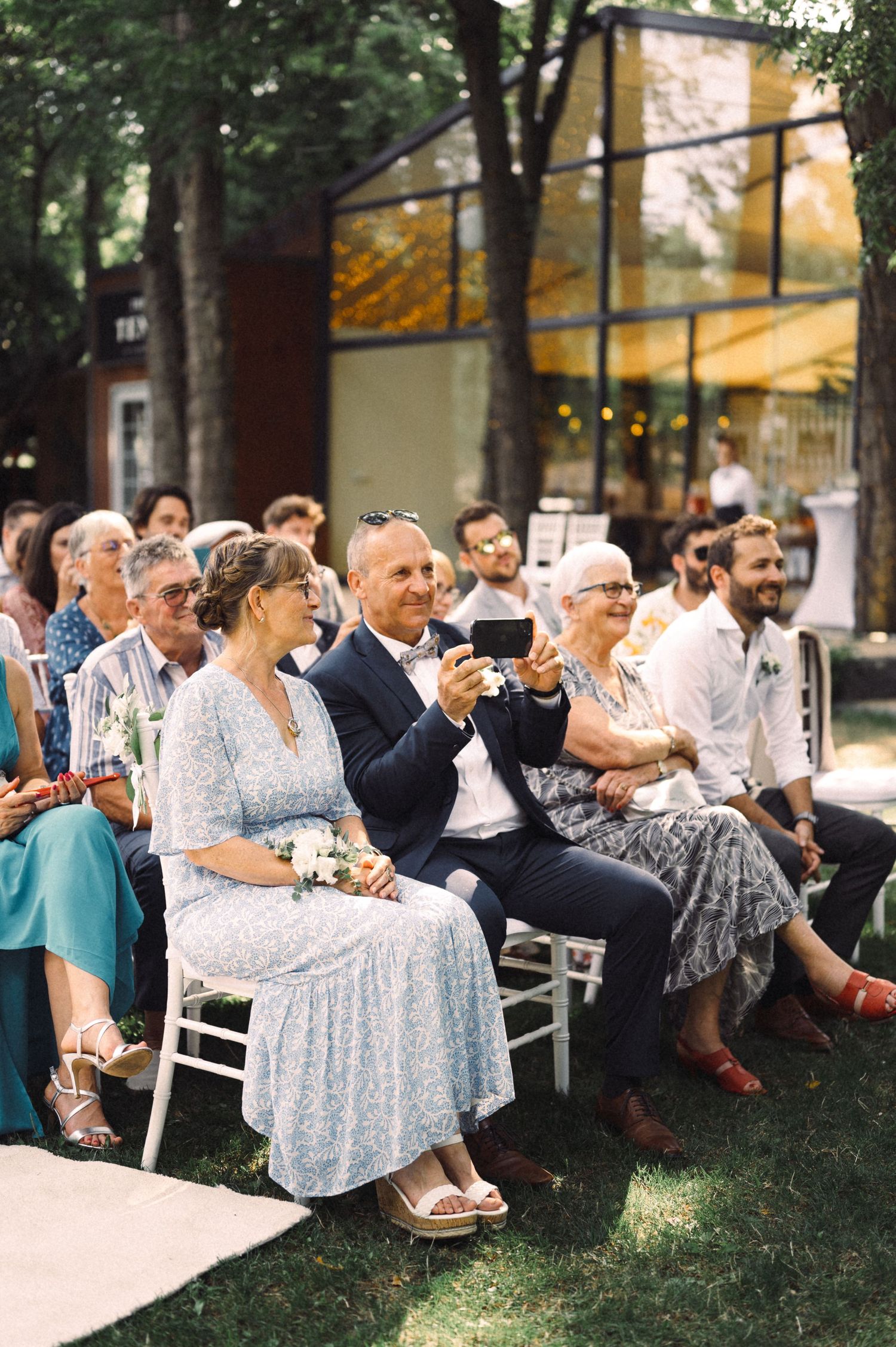 Wedding guests seated on white chairs enjoying the outdoor ceremony while taking photos.