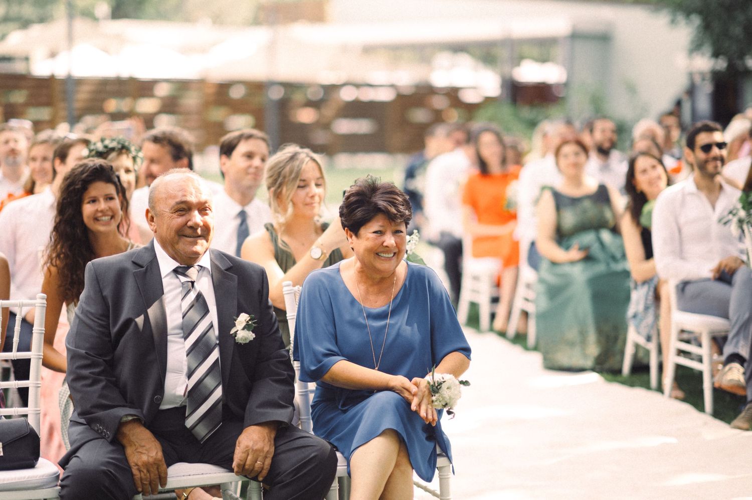Wedding guests seated in formal attire along white aisle runner during outdoor ceremony.