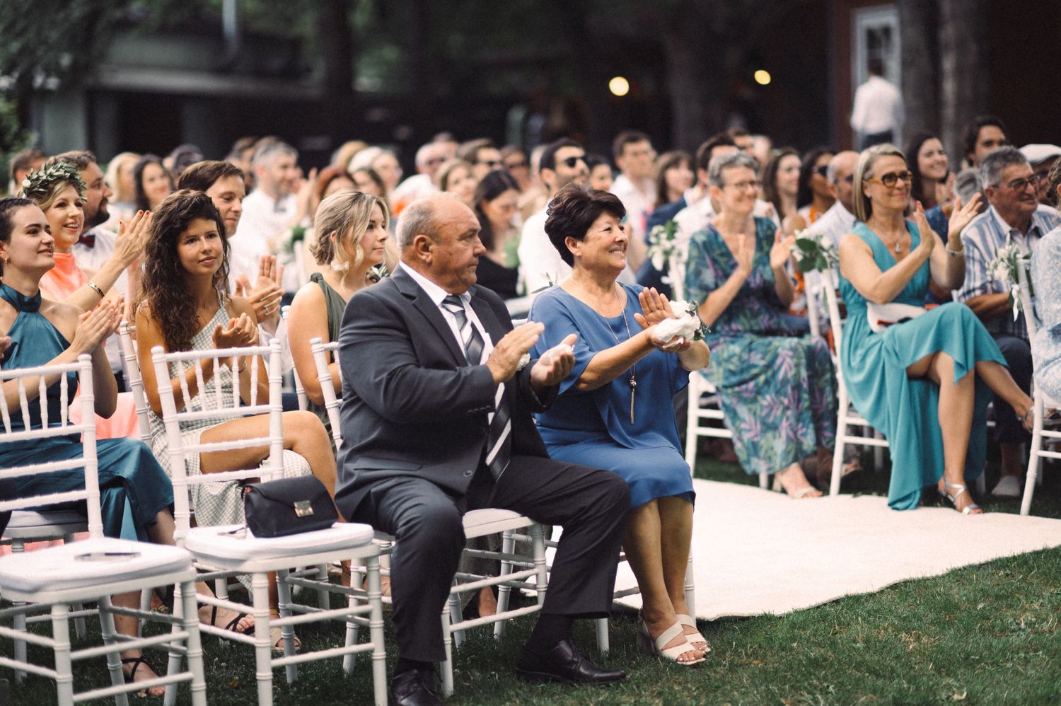 Wedding guests applauding during an outdoor ceremony with white chairs.