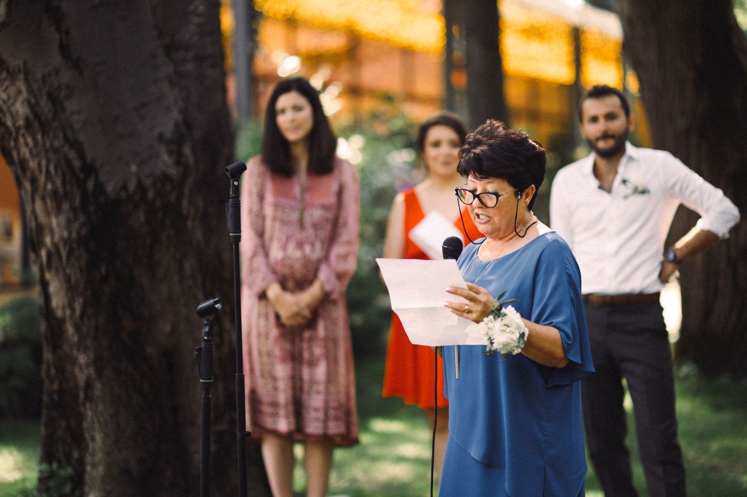 A young person in blue reads from a paper during an outdoor ceremony while others watch in the background.