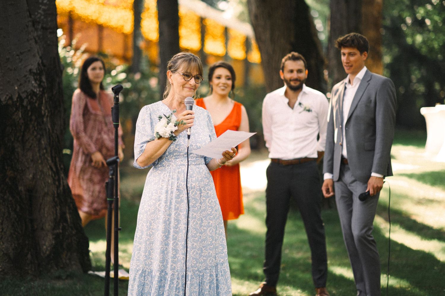 Person in a floral dress delivers a speech at an outdoor garden event with attendees looking on.