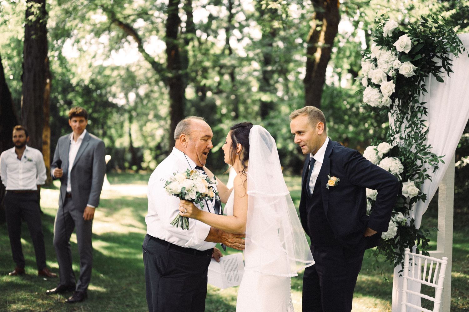 Outdoor wedding ceremony under trees with white floral arch decorations.