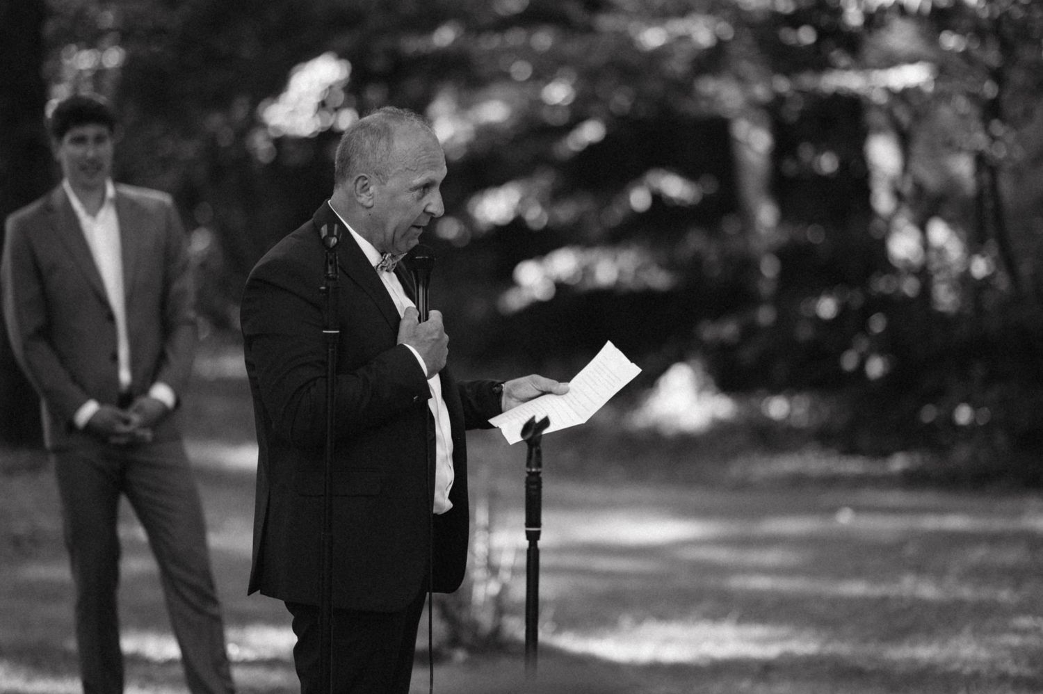Black and white photo of speaker reading from paper during outdoor ceremony.