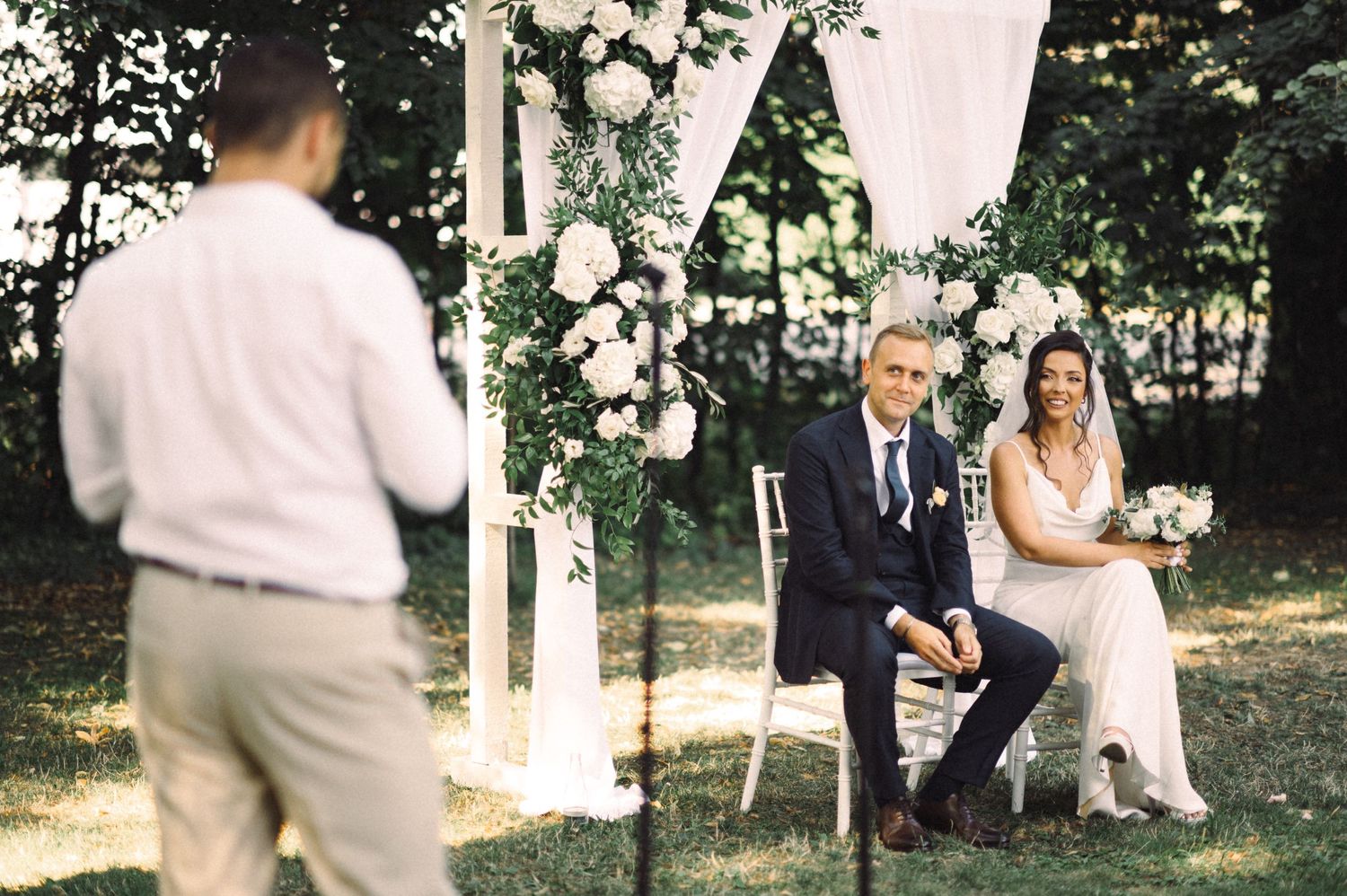 Outdoor wedding ceremony setup with white arch and floral decorations in a wooded setting.