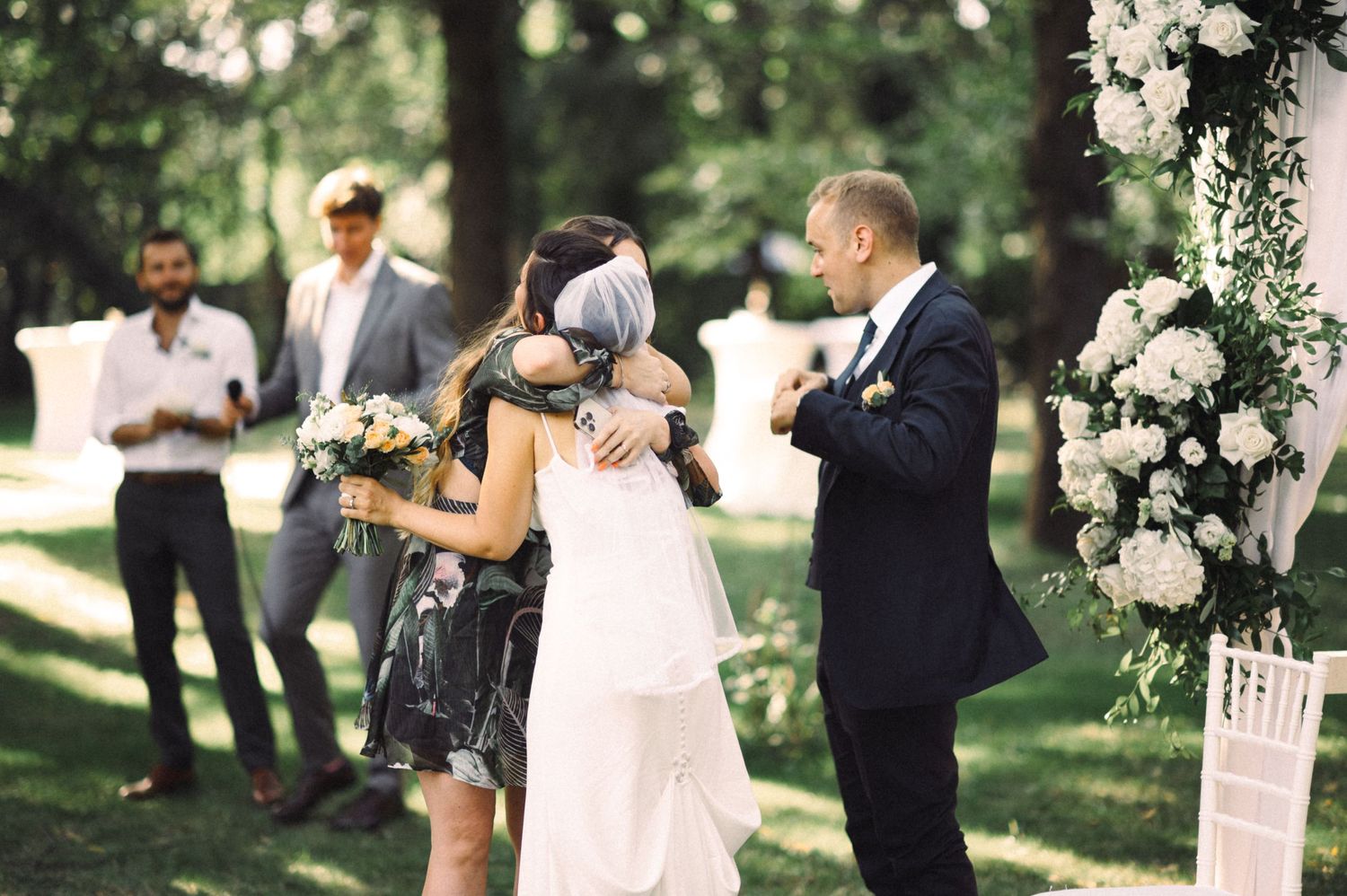 Guests celebrate during an outdoor wedding ceremony under white floral decorations in a garden setting.
