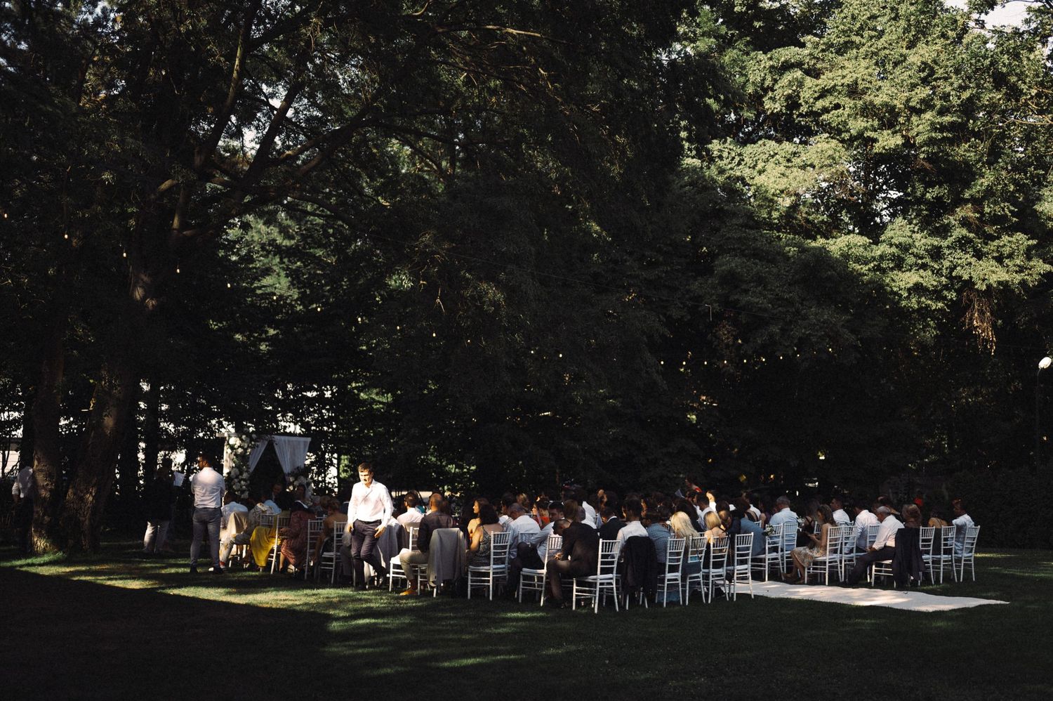 White chairs arranged in rows for an outdoor wedding ceremony under shady trees.