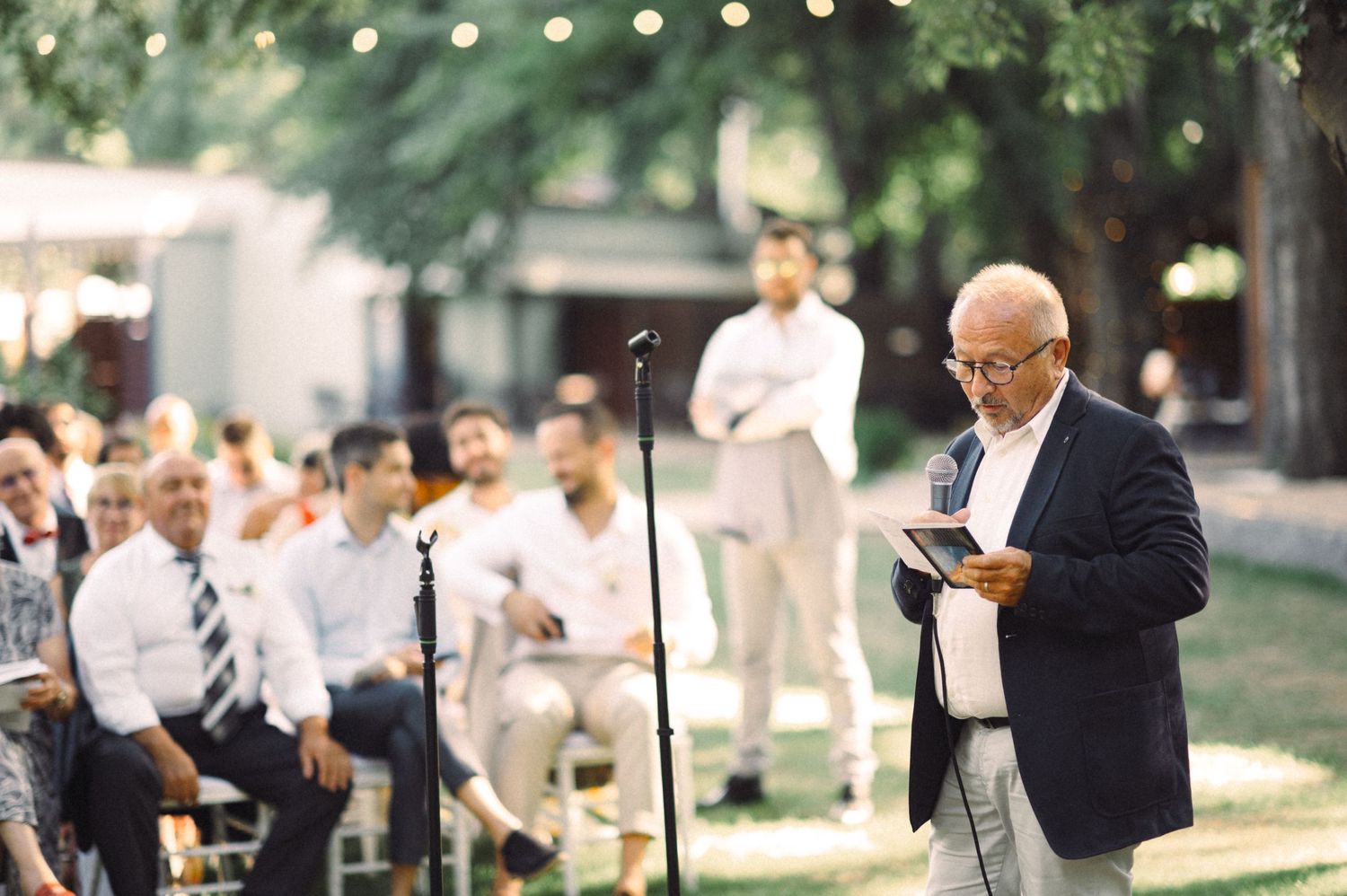 Speaker addresses seated guests at outdoor ceremony with string lights overhead.