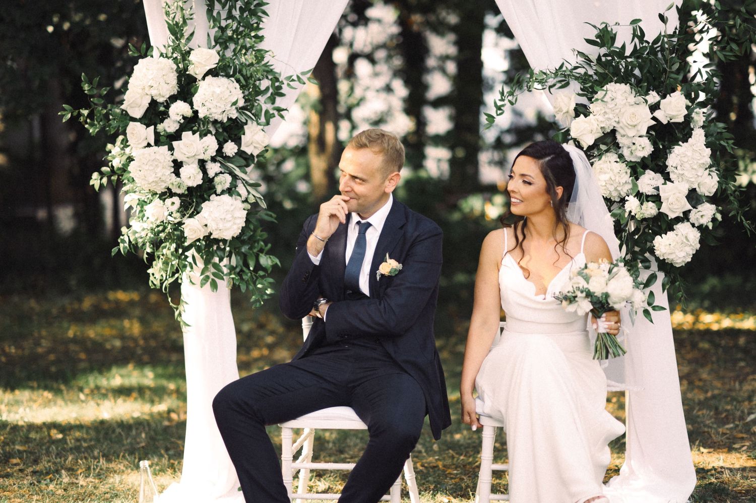 Couple seated under white floral arch during outdoor garden ceremony.