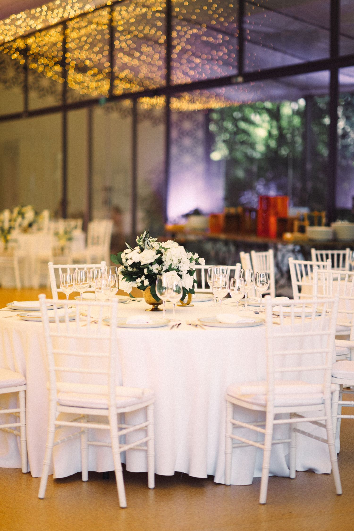 Wedding reception venue with white chiavari chairs and tables under twinkling lights and greenery.