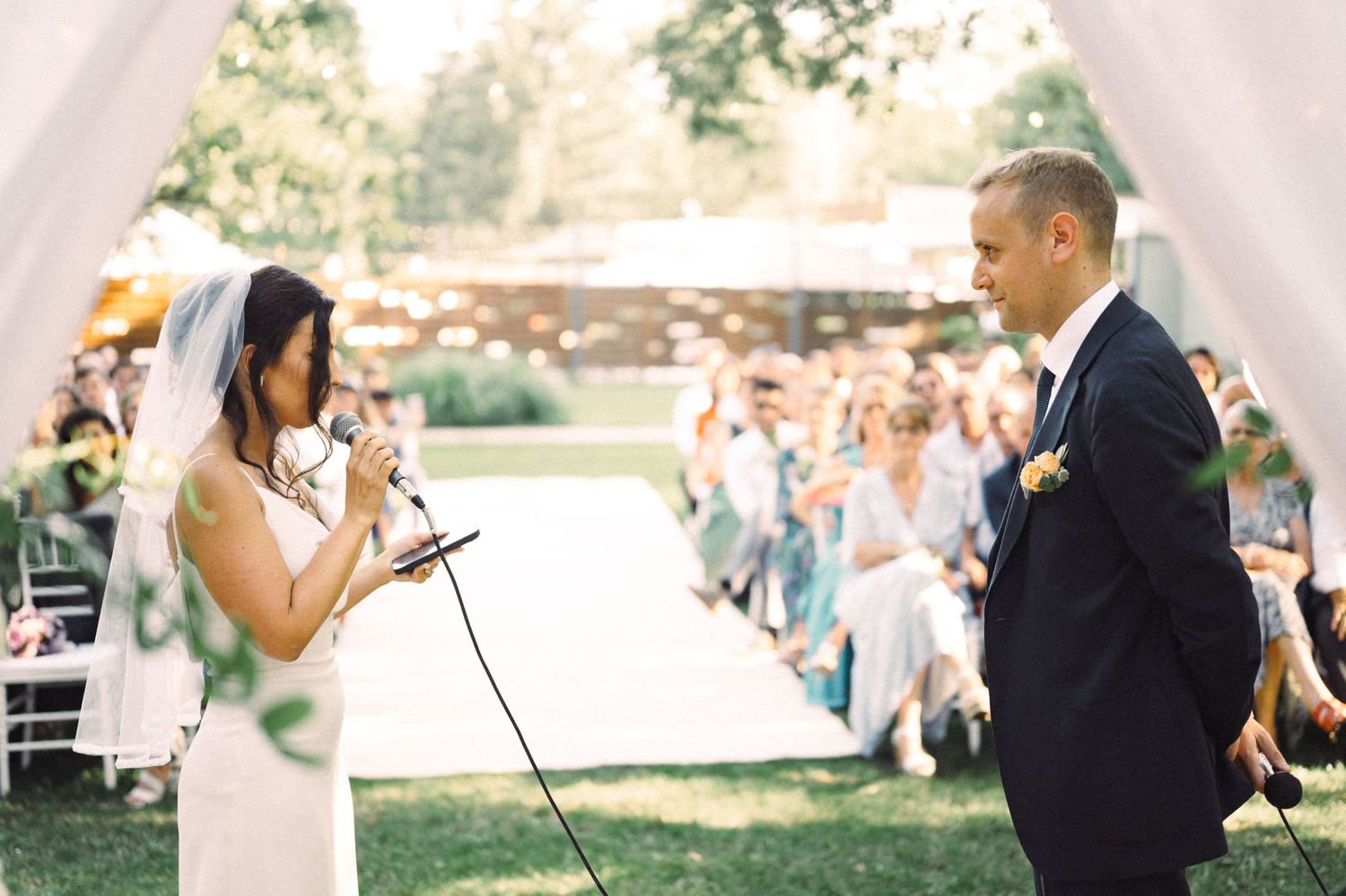 Outdoor wedding ceremony with guests watching couple exchange vows under white decorative arch.