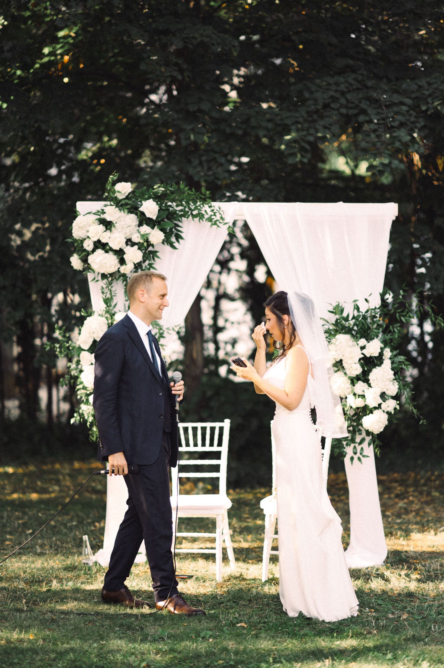 Outdoor wedding ceremony with white floral arch and elegant decor in a garden setting.