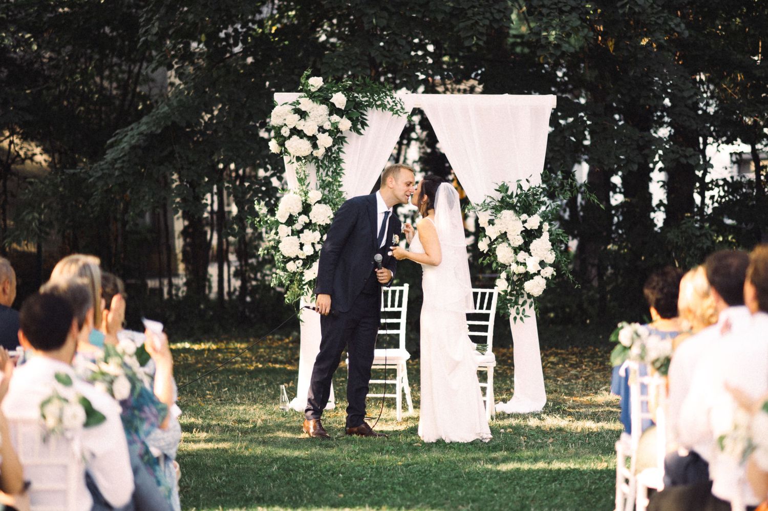Garden wedding ceremony with white floral arch and seated guests on a sunny afternoon.