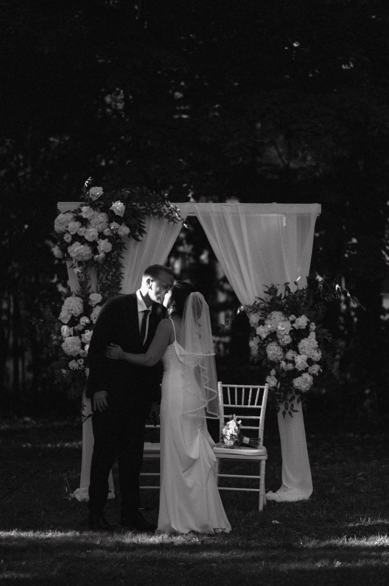 A black and white photo of a wedding ceremony under an elegant floral arch with flowing white drapes.