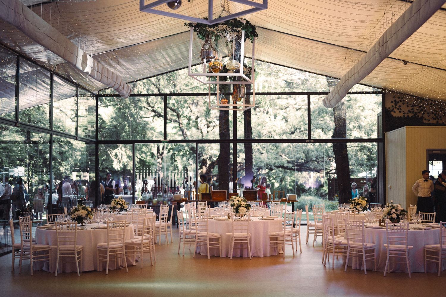 A spacious wedding reception venue with pink tablecloths and elegant chandeliers under a glass ceiling.