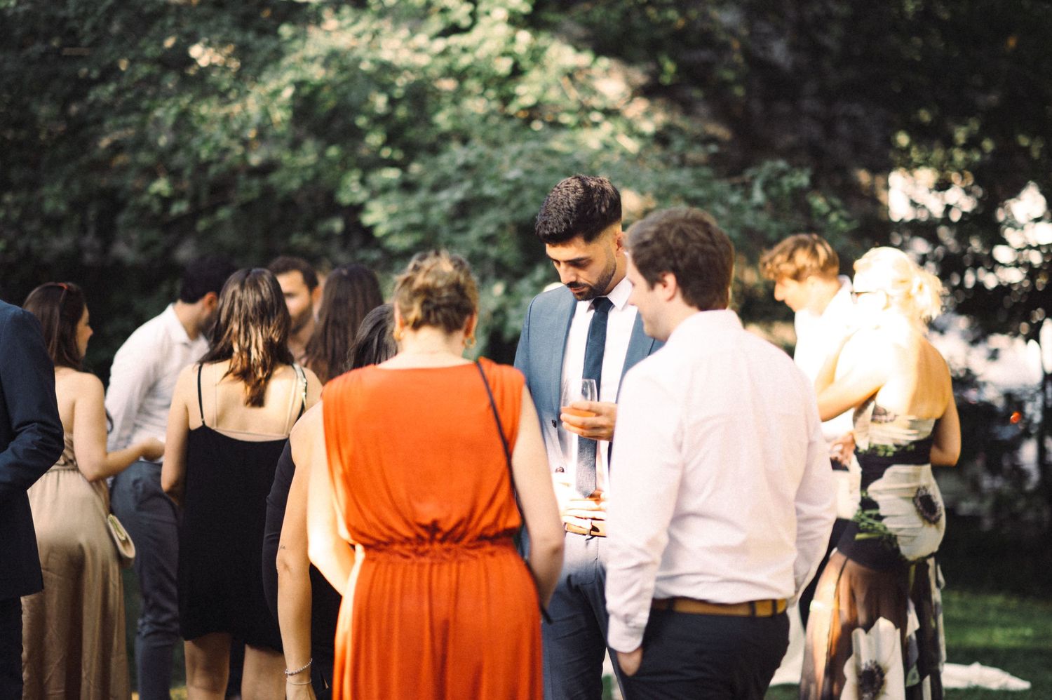 Wedding guests mingling at an outdoor cocktail reception with someone wearing a vibrant orange dress.