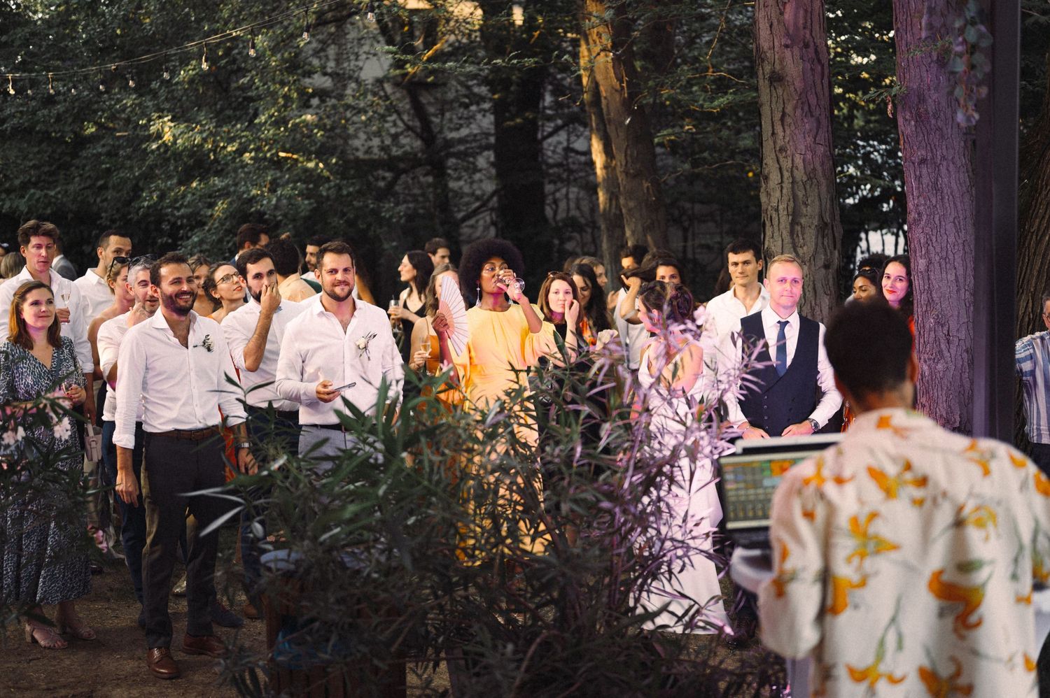 Large group of celebration attendees gathered under trees with string lights at dusk.