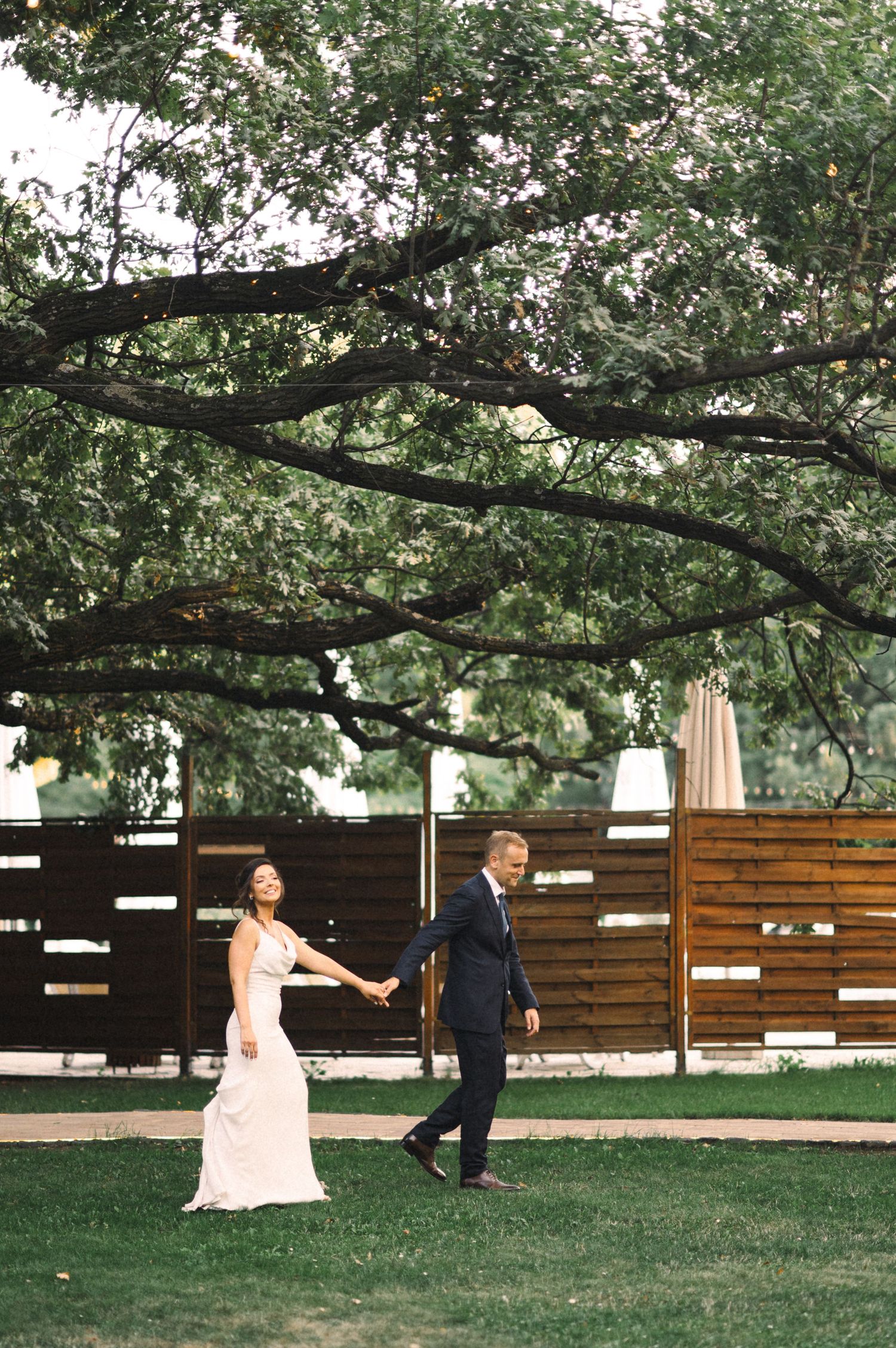 Wedding couple dances playfully on a lawn beneath sprawling tree branches.
