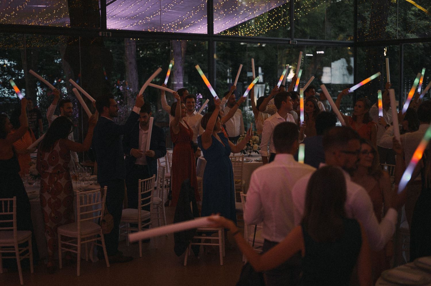 Wedding reception party with guests waving light sticks under a tent at night.