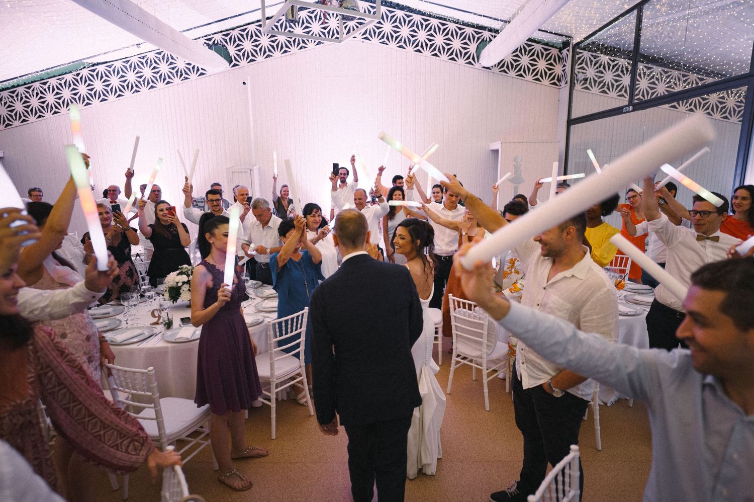 Wedding reception guests raise their arms in celebration inside a bright white venue with elegant table settings.